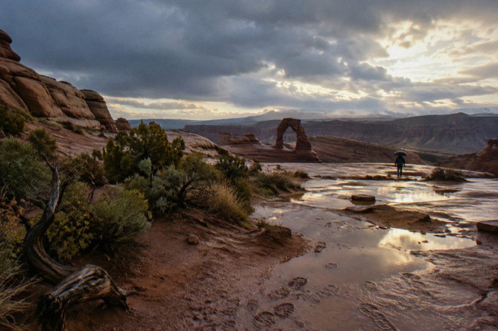 Storm breaking over distant arch, wet slickrock reflecting clouds, boot-printed mud, figure with umbrella