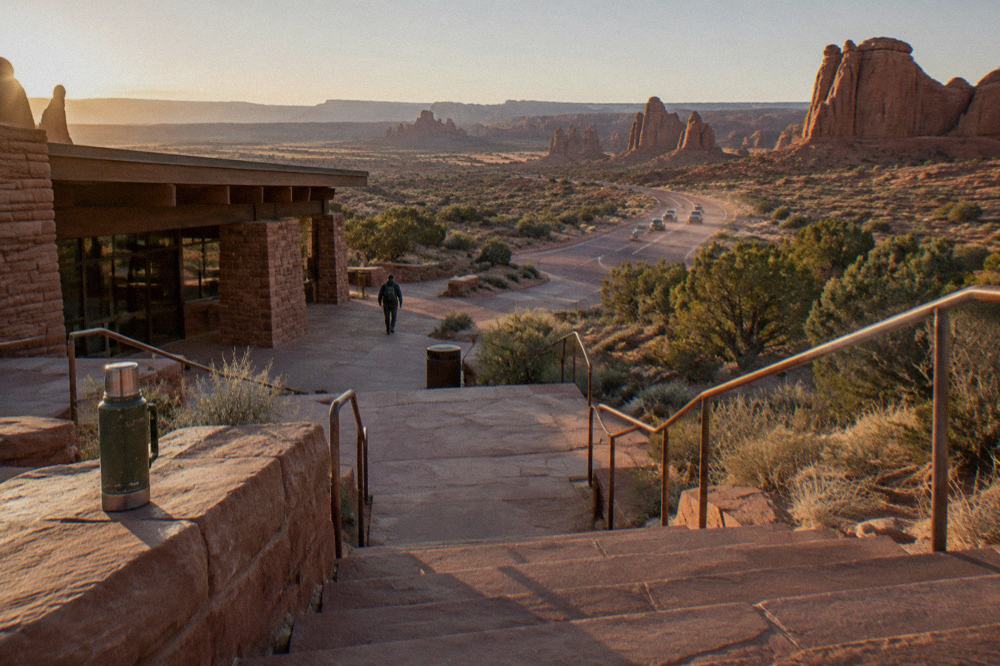 Morning sun on sandstone switchback steps, three cars climbing entrance road, heat haze, thermos on steps