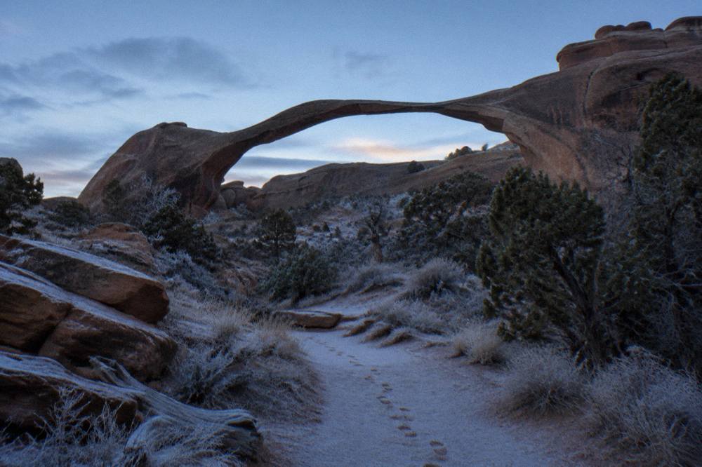 A long thin sandstone arch spanning a quiet desert ridge with no people on the trail below it
