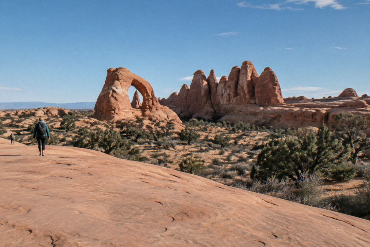 Red sandstone fins and a lone sandstone arch rising from empty desert scrubland under a wide blue sky