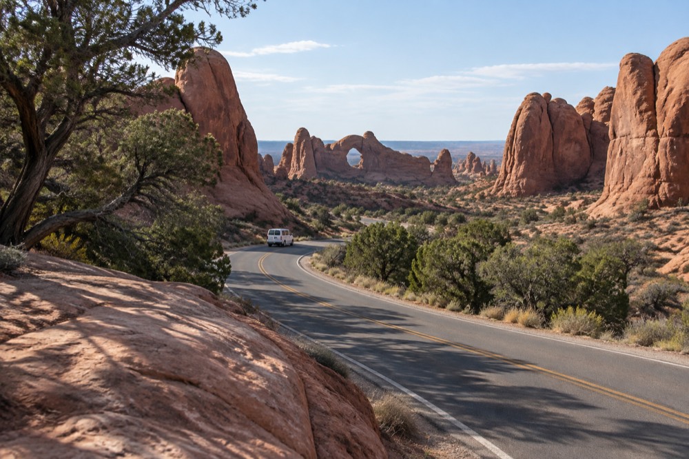 A paved park road curving between tall sandstone fins under a wide blue sky