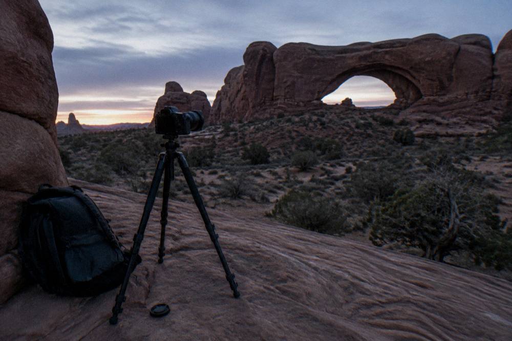 Tripod and camera bag set on slickrock facing a wide arch opening before dawn