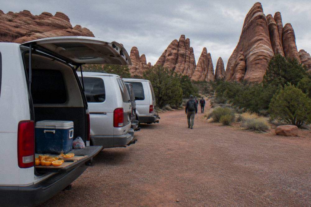 A row of tour vans parked at a trailhead pullout beside tall sandstone fins