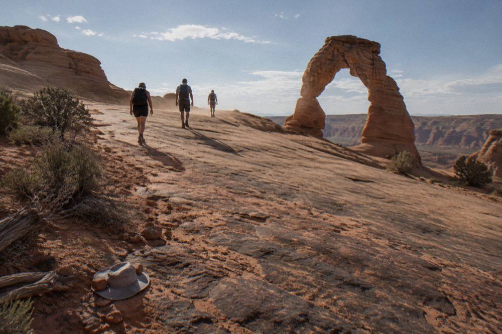 Hikers on a broad slickrock slope below Delicate Arch in late afternoon light