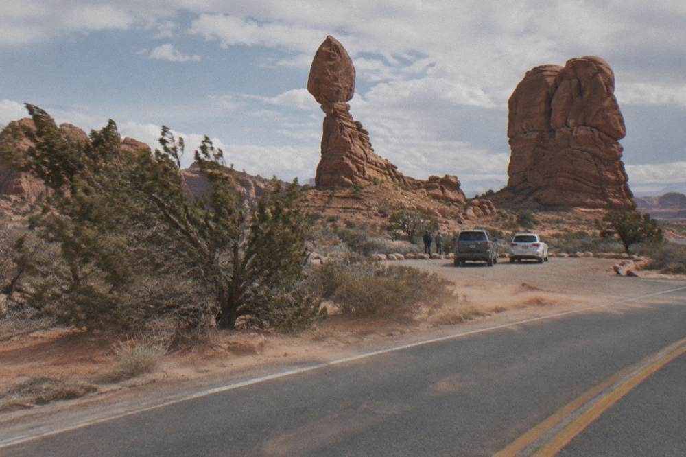 A paved park road curving between tall red sandstone formations with cars parked at a viewpoint.