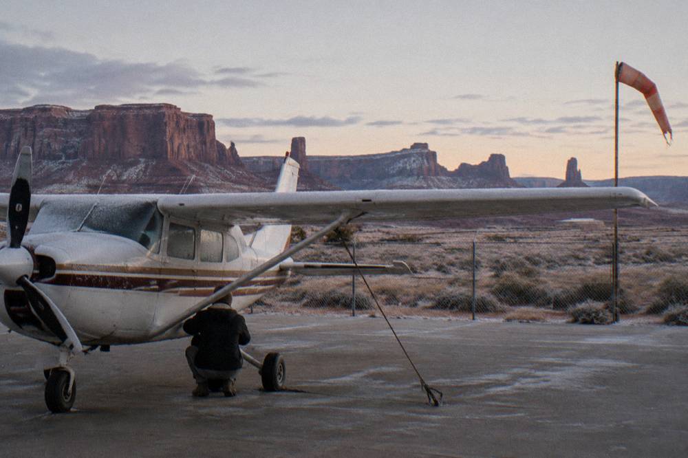 A high-wing airplane parked on a desert airstrip with red cliffs in the background.