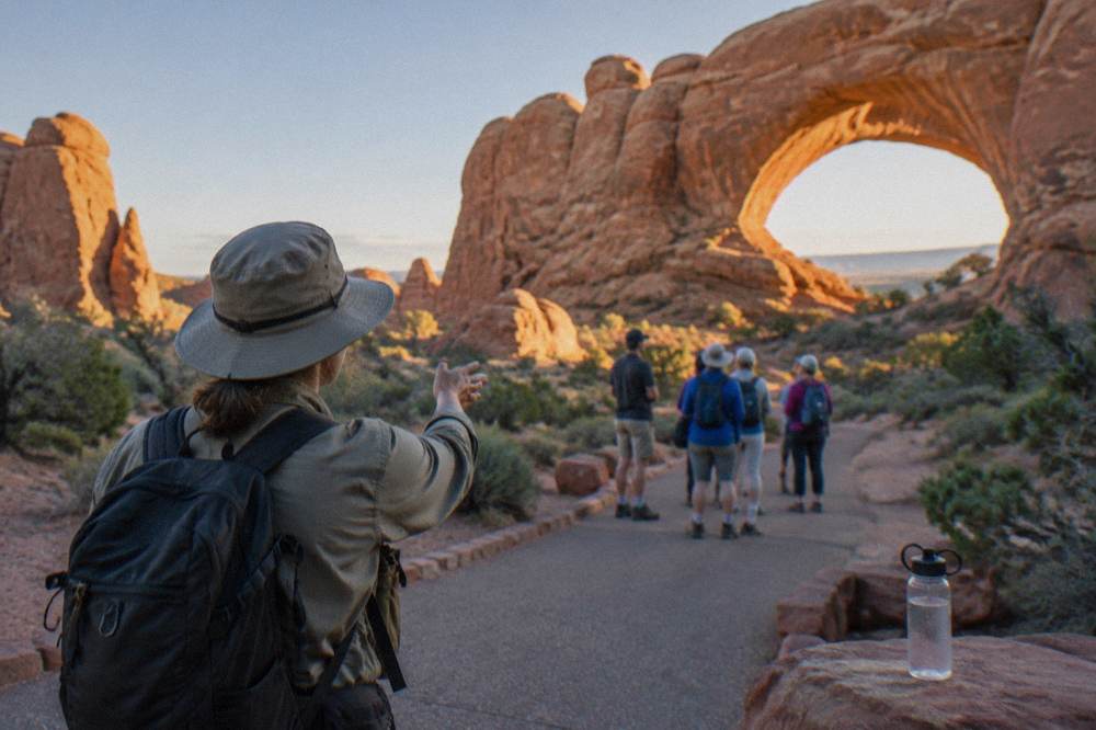 A guide gesturing toward a sandstone arch while a small group listens on a paved trail