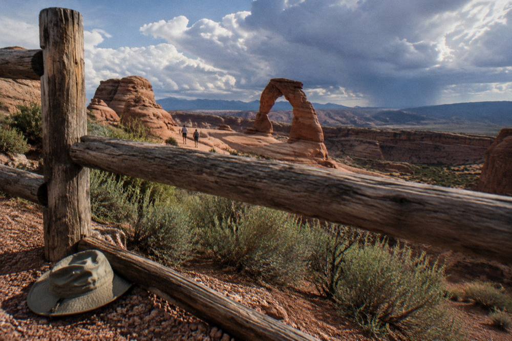 A large sandstone arch framed by a viewpoint railing under scattered clouds