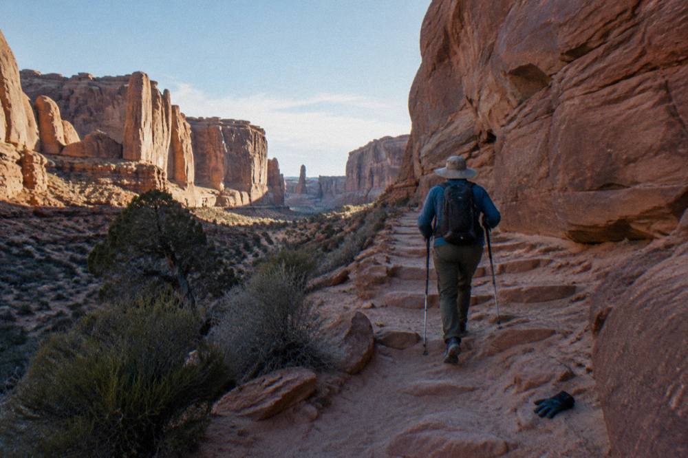 A hiker in a sun hat and long sleeves walking a sandy trail between tall red rock walls