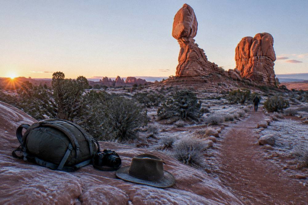 A packed daypack on a rock ledge with a water bottle, hat and camera at sunrise