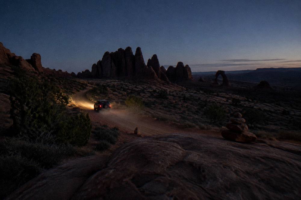 Lone vehicle on a desert road at dawn heading toward sandstone fins